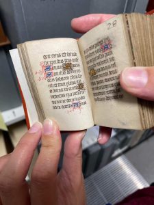 Small medieval manuscript held open to show Latin prayer text, written in black ink with illuminated initials.