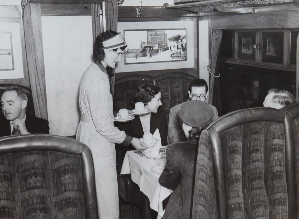 Black and white photograph of a woman serving tea to a group in a train carriage booth.