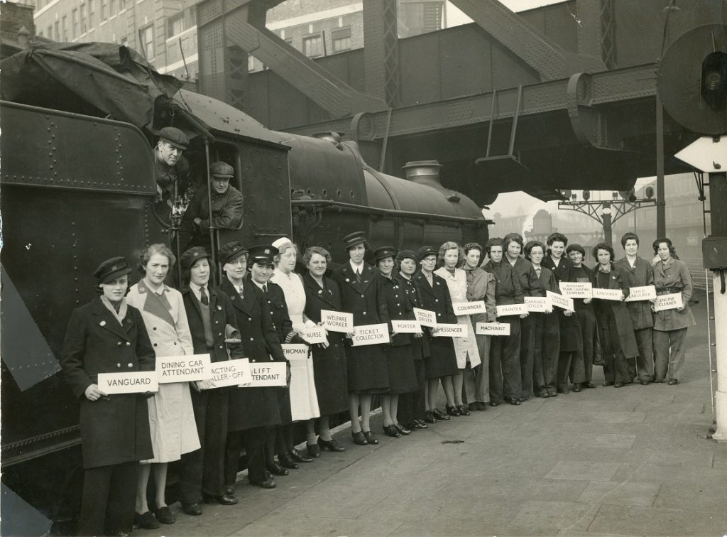 Black and white photograph of women railway workers during WWII.  Each woman holds a board stating her job title - van guard, dining car attendant, ticket collector, porter etc. 