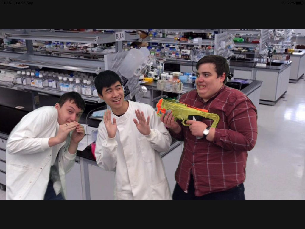 Two students wearing lab coats stand in a lab, with a student pointing a plastic green gun at them.