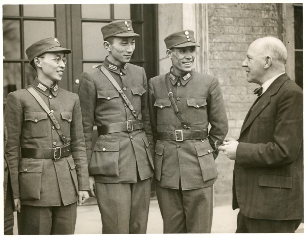 Black and white photograph of 3 Chinese students in uniform. They are standing outside a building talking to F. L. Attenborough, Principal of University College Leicester, 1945